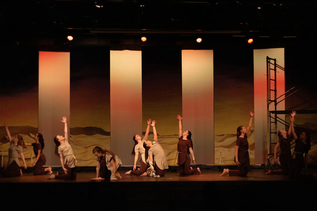 An ensemble kneeling before a desert backdrop and tall stage panels.
