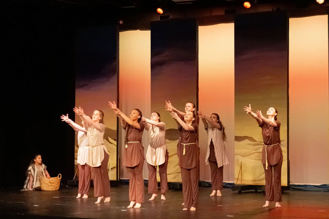 Dancers in earth-toned costumes reaching forward onstage before a desert backdrop.