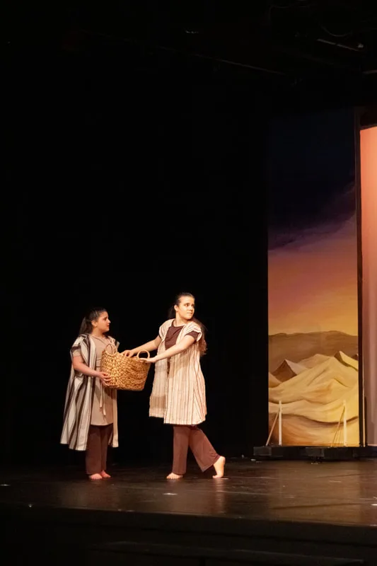 Two dancers carrying a basket beside the desert stage set.