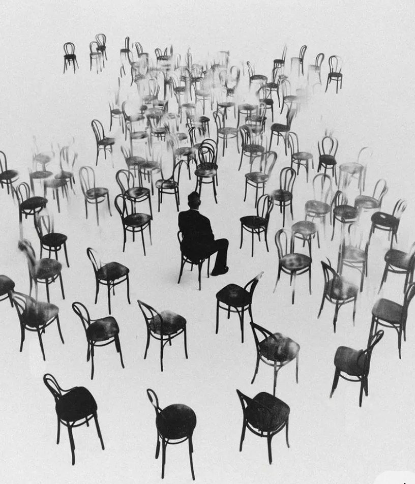 Dozens of black bentwood chairs scattered across a white space, one figure seated among them.