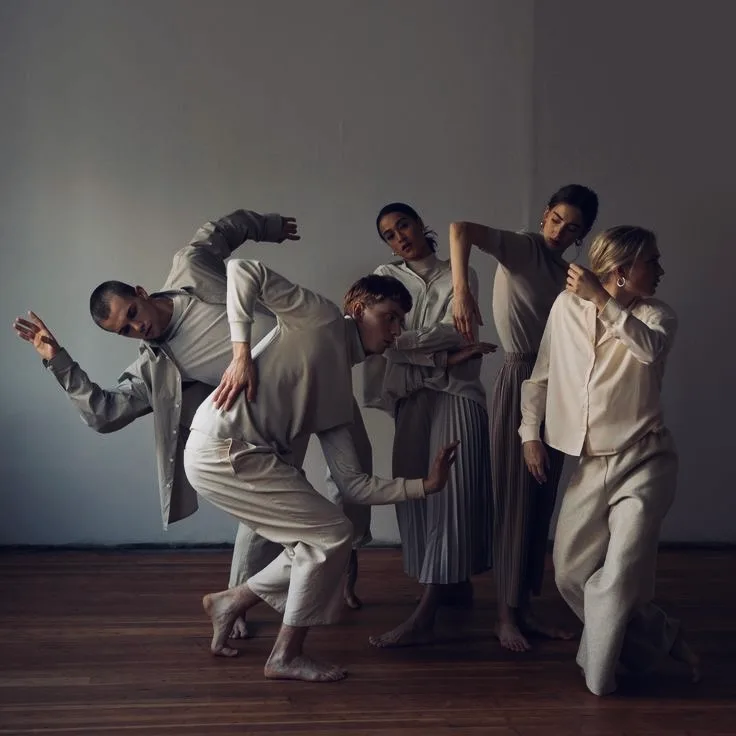 A group of dancers in cream clothing leaning into one another to form a single shape.