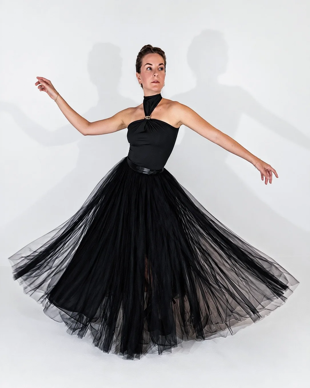 A woman in a black halter top and full black tulle skirt, mid-turn against a pale backdrop.