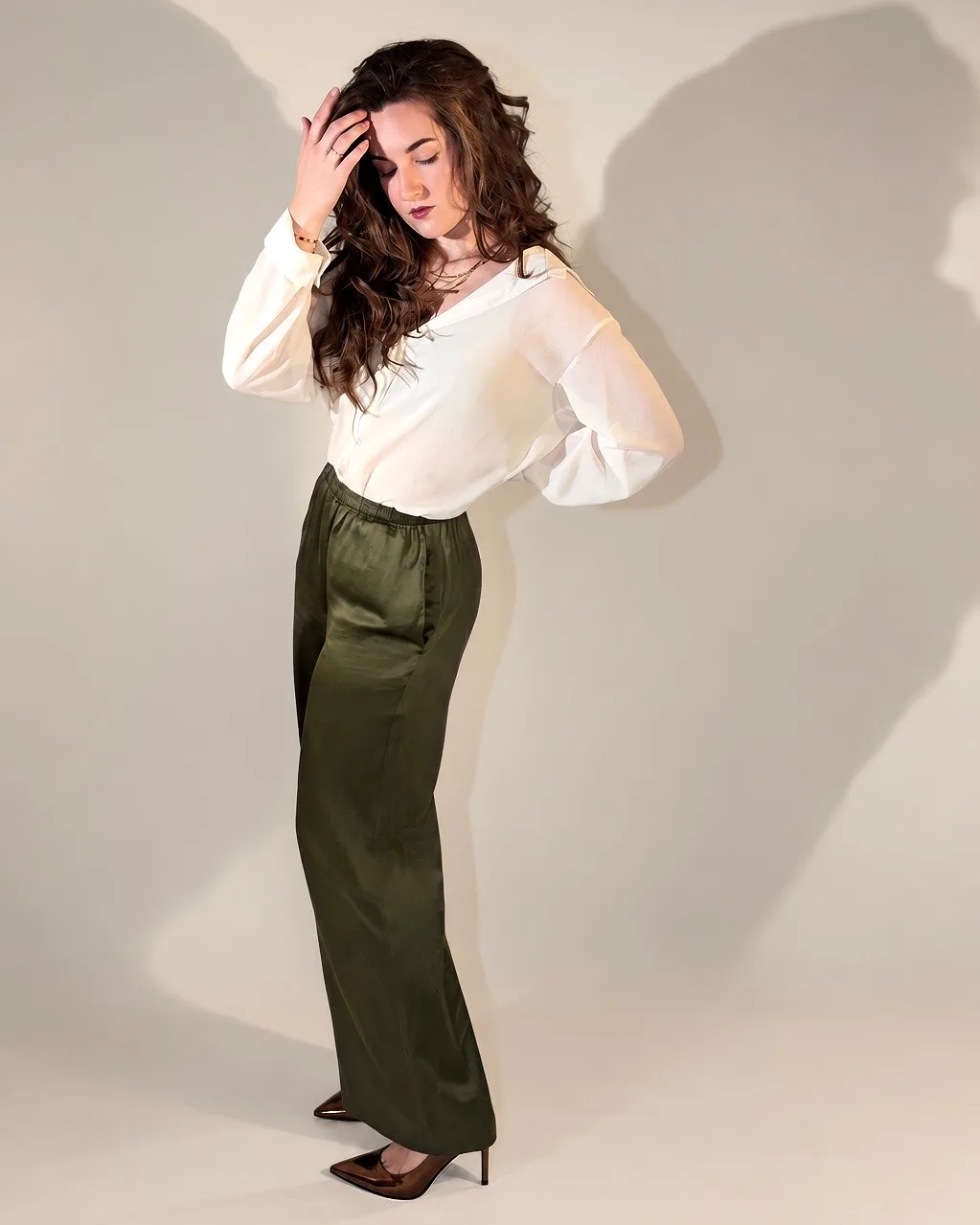 A woman in a white blouse and olive satin trousers, head tilted down, one hand in her hair, in warm light.