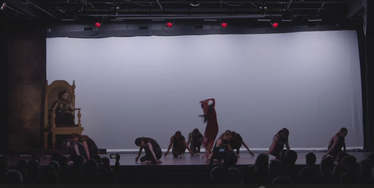 Dancers crouched low before a pale lavender backdrop, with the throne at stage left.
