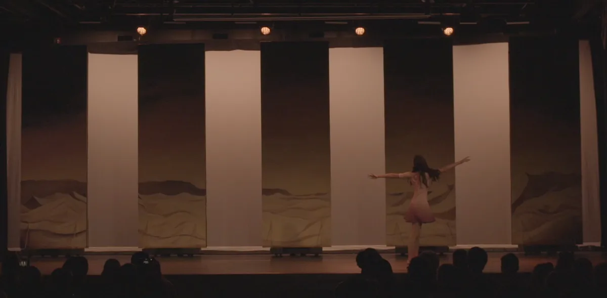 A solo dancer mid-leap before tall pale panels and a desert backdrop.